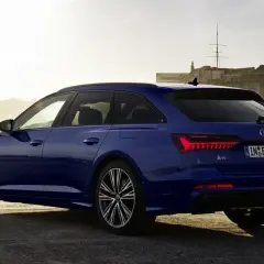 Niebieskie Audi A6 Avant z napisem "50 TFSI e" na klapie bagażnika, zaparkowane przy nabrzeżu z widokiem na zabytkową fortecę i żaglówki.