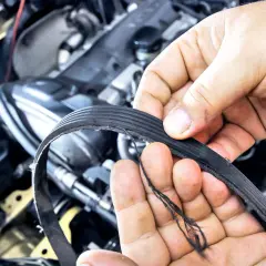 Zerwany pasek alternatora, z widocznymi poszarpanymi włóknami, trzymany w dłoniach mechanika na tle silnika samochodowego.