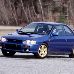Niebieskie Subaru Impreza z lat 90. na złotych felgach, z kierowcą za kierownicą, przemierza pustą drogę. To auto, które kojarzy się z rajdami i Japonią.