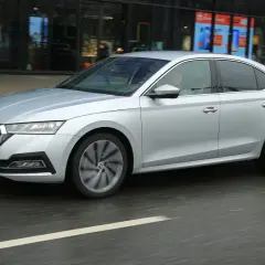 Srebrny sedan z napędem 1.5 TSI mHEV porusza się po miejskiej ulicy, mijając witryny sklepowe.