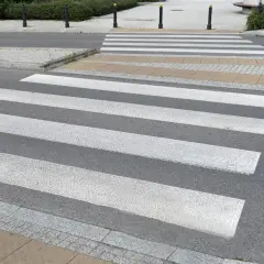 Sugerowane przejście dla pieszych z białymi pasami na szarym asfalcie, prowadzące przez chodnik i trawnik.