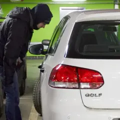 Mężczyzna w kapturze pochyla się nad białym Volkswagenem Golfem, jednym z najczęściej kradzionych aut.