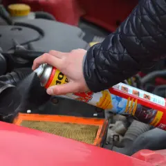 Ręka trzyma puszkę z preparatem do odpalania na plaka, rozpylając go w okolicy filtra powietrza silnika.