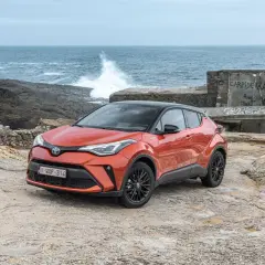 Pomarańczowe, małe auta hybrydowe Toyota C-HR na kamienistym wybrzeżu. W tle widać morze z falami i zniszczony budynek.