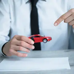 Mężczyzna w białej koszuli i czarnej krawacie prezentuje czerwony model samochodu, wskazując na niego palcem. Obok leży dokument, sugerujący umowę kupna sprzedaży samochodu z wyłączeniem rękojmi.