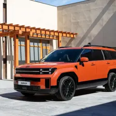 Pomarańczowy SUV Hyundai Santa Fe z czarnymi dodatkami i felgami, zaparkowany przed budynkiem z drewnianą pergolą.
