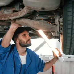 Mechanik w niebieskim kombinezonie oświetla latarką podwozie samochodu. Zastanawiasz się, ile trwa przegląd samochodu?