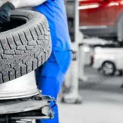 Mechanik w niebieskim kombinezonie trzyma oponę zimową. W tle widać podnośnik samochodowy i czerwony samochód. Sprawdź aktualne ceny wymiany opon.