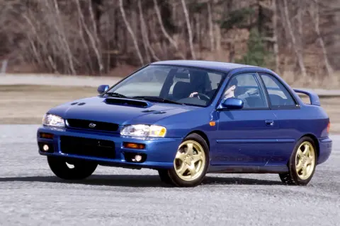 Niebieskie Subaru Impreza z lat 90. na złotych felgach, z kierowcą za kierownicą, przemierza pustą drogę. To auto, które kojarzy się z rajdami i Japonią.