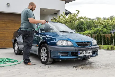 Mężczyzna myje niebieski samochód na posesji, używając węża ogrodowego. Woda spływa po karoserii i tworzy kałuże na podjeździe.
