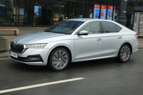 Srebrny sedan z napędem 1.5 TSI mHEV porusza się po miejskiej ulicy, mijając witryny sklepowe.