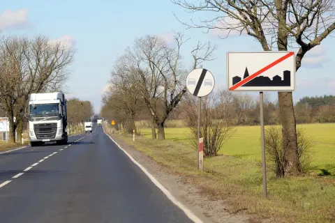 Znaki drogowe informują, ile można jechać poza terenem zabudowanym. Ciężarówka jedzie drogą, mijając pola i drzewa.