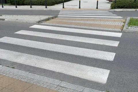 Sugerowane przejście dla pieszych z białymi pasami na szarym asfalcie, prowadzące przez chodnik i trawnik.