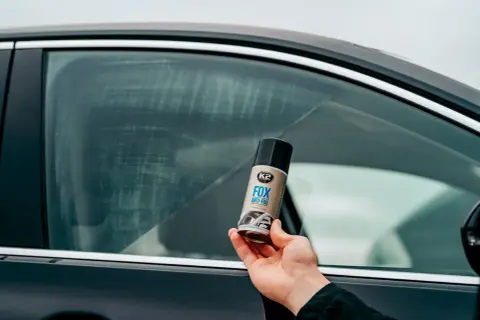 Dłoń trzyma puszkę K2 FOX ANTI-FOG, która pomoże Ci jak odparować szyby w aucie. Widoczne smugi na szybie sugerują problem z zaparowaniem.