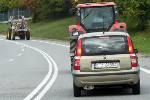 Złoty Fiat Panda jedzie za czerwonym traktorem. Czy można wyprzedzić traktor na podwójnej ciągłej? Droga kręta, drzewa po bokach.