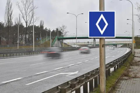 Znak drogowy: biały romb na niebieskim tle. Droga szybkiego ruchu z rozmytymi samochodami i zielonym mostem dla pieszych.