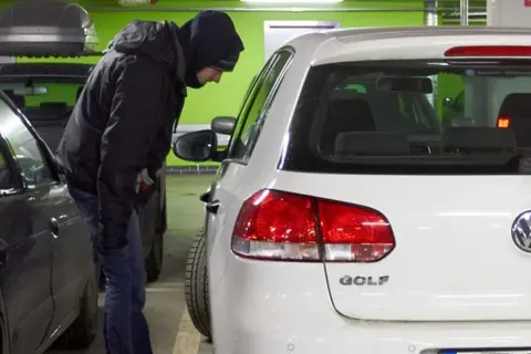 Mężczyzna w kapturze pochyla się nad białym Volkswagenem Golfem, jednym z najczęściej kradzionych aut.