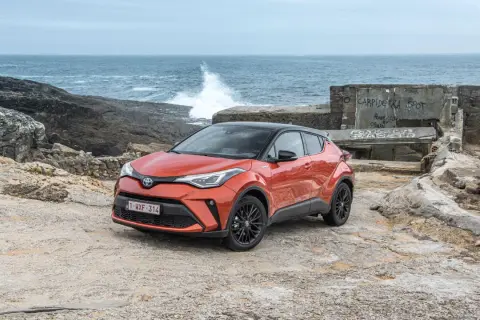 Pomarańczowe, małe auta hybrydowe Toyota C-HR na kamienistym wybrzeżu. W tle widać morze z falami i zniszczony budynek.