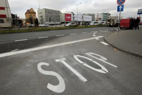 Na asfalcie widoczna jest biała linia warunkowego zatrzymania "STOP". W tle budynki i pojazdy.