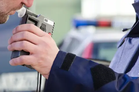 Mężczyzna dmucha w alkomat, który pokazuje wynik, mimo że nie pił. Policjant w mundurze z napisem POLICJA trzyma urządzenie.