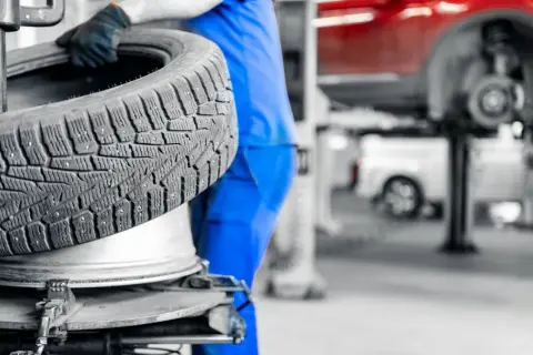 Mechanik w niebieskim kombinezonie trzyma oponę zimową. W tle widać podnośnik samochodowy i czerwony samochód. Sprawdź aktualne ceny wymiany opon.