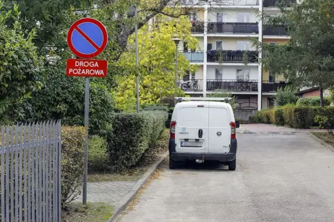 Znak zakazu postoju na drodze pożarowej. Biały bus stoi na poboczu, gdzie nie można parkować.