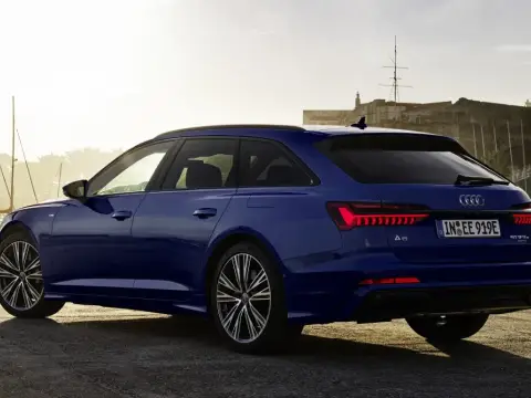 Niebieskie Audi A6 Avant z napisem "50 TFSI e" na klapie bagażnika, zaparkowane przy nabrzeżu z widokiem na zabytkową fortecę i żaglówki.