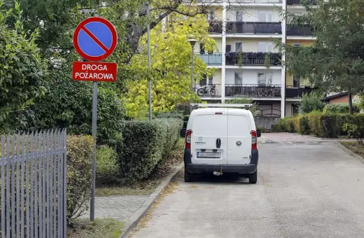 Znak zakazu postoju na drodze pożarowej. Biały bus stoi na poboczu, gdzie nie można parkować.