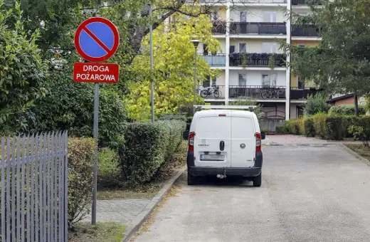 Znak zakazu postoju na drodze pożarowej. Biały bus stoi na poboczu, gdzie nie można parkować.