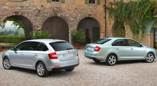 Dwa samochody: srebrny liftback i błękitny hatchback, stoją przed kamiennym budynkiem z łukami i zielenią.