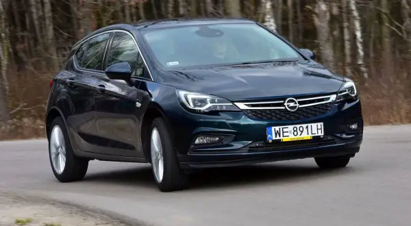 Ciemnozielona Astra K 1.6 CDTI w ruchu, z widocznym kierowcą za kierownicą, na tle lasu.