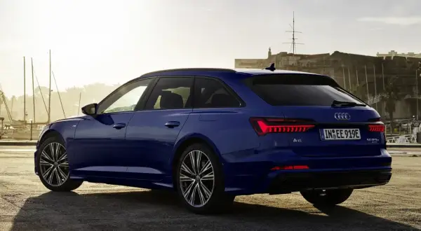 Niebieskie Audi A6 Avant z napisem "50 TFSI e" na klapie bagażnika, zaparkowane przy nabrzeżu z widokiem na zabytkową fortecę i żaglówki.
