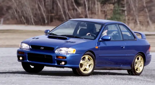 Niebieskie Subaru Impreza z lat 90. na złotych felgach, z kierowcą za kierownicą, przemierza pustą drogę. To auto, które kojarzy się z rajdami i Japonią.