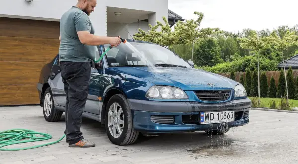 Mężczyzna myje niebieski samochód na posesji, używając węża ogrodowego. Woda spływa po karoserii i tworzy kałuże na podjeździe.