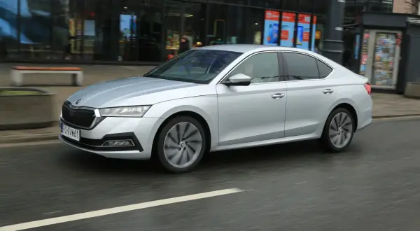 Srebrny sedan z napędem 1.5 TSI mHEV porusza się po miejskiej ulicy, mijając witryny sklepowe.