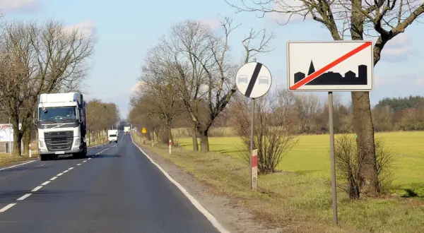 Znaki drogowe informują, ile można jechać poza terenem zabudowanym. Ciężarówka jedzie drogą, mijając pola i drzewa.