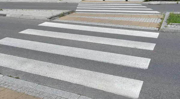 Sugerowane przejście dla pieszych z białymi pasami na szarym asfalcie, prowadzące przez chodnik i trawnik.