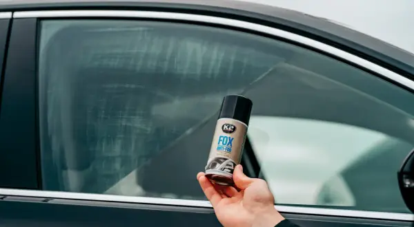 Dłoń trzyma puszkę K2 FOX ANTI-FOG, która pomoże Ci jak odparować szyby w aucie. Widoczne smugi na szybie sugerują problem z zaparowaniem.