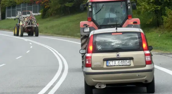 Złoty Fiat Panda jedzie za czerwonym traktorem. Czy można wyprzedzić traktor na podwójnej ciągłej? Droga kręta, drzewa po bokach.