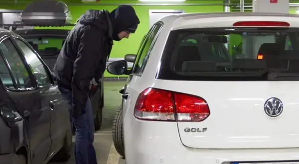 Mężczyzna w kapturze pochyla się nad białym Volkswagenem Golfem, jednym z najczęściej kradzionych aut.