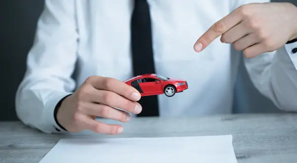 Mężczyzna w białej koszuli i czarnej krawacie prezentuje czerwony model samochodu, wskazując na niego palcem. Obok leży dokument, sugerujący umowę kupna sprzedaży samochodu z wyłączeniem rękojmi.