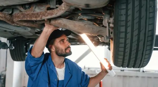 Mechanik w niebieskim kombinezonie oświetla latarką podwozie samochodu. Zastanawiasz się, ile trwa przegląd samochodu?