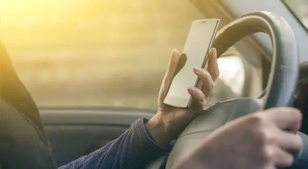 Ręka trzymająca telefon podczas jazdy samochodem. Słońce świeci przez szybę, oślepiając kierowcę.