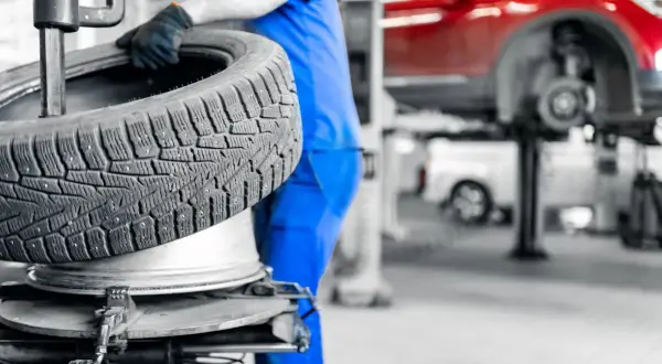 Mechanik w niebieskim kombinezonie trzyma oponę zimową. W tle widać podnośnik samochodowy i czerwony samochód. Sprawdź aktualne ceny wymiany opon.