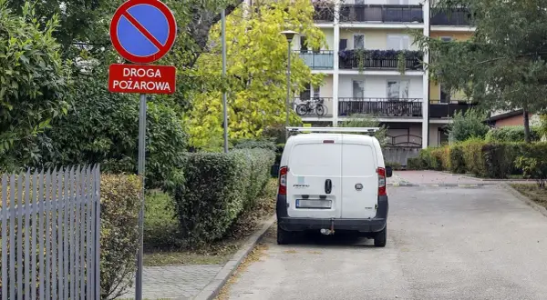 Znak zakazu postoju na drodze pożarowej. Biały bus stoi na poboczu, gdzie nie można parkować.
