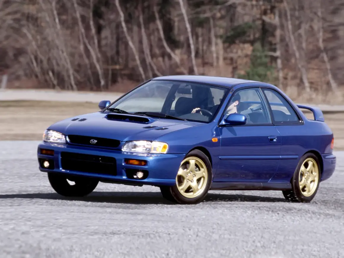 Niebieskie Subaru Impreza z lat 90. na złotych felgach, z kierowcą za kierownicą, przemierza pustą drogę. To auto, które kojarzy się z rajdami i Japonią.