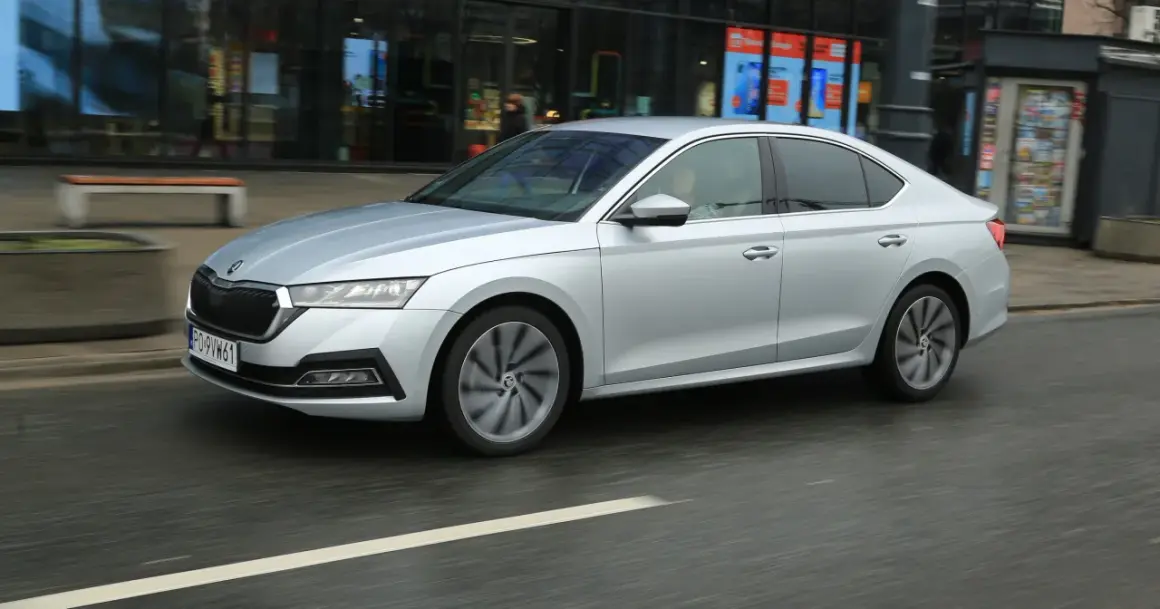 Srebrny sedan z napędem 1.5 TSI mHEV porusza się po miejskiej ulicy, mijając witryny sklepowe.