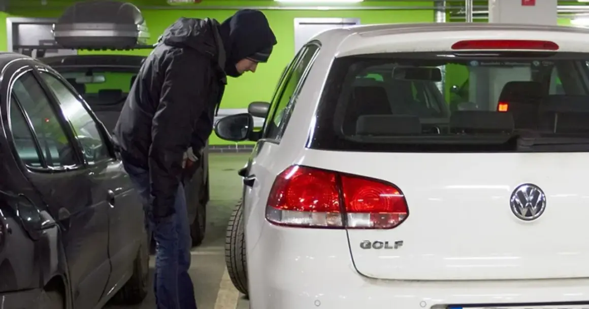 Mężczyzna w kapturze pochyla się nad białym Volkswagenem Golfem, jednym z najczęściej kradzionych aut.