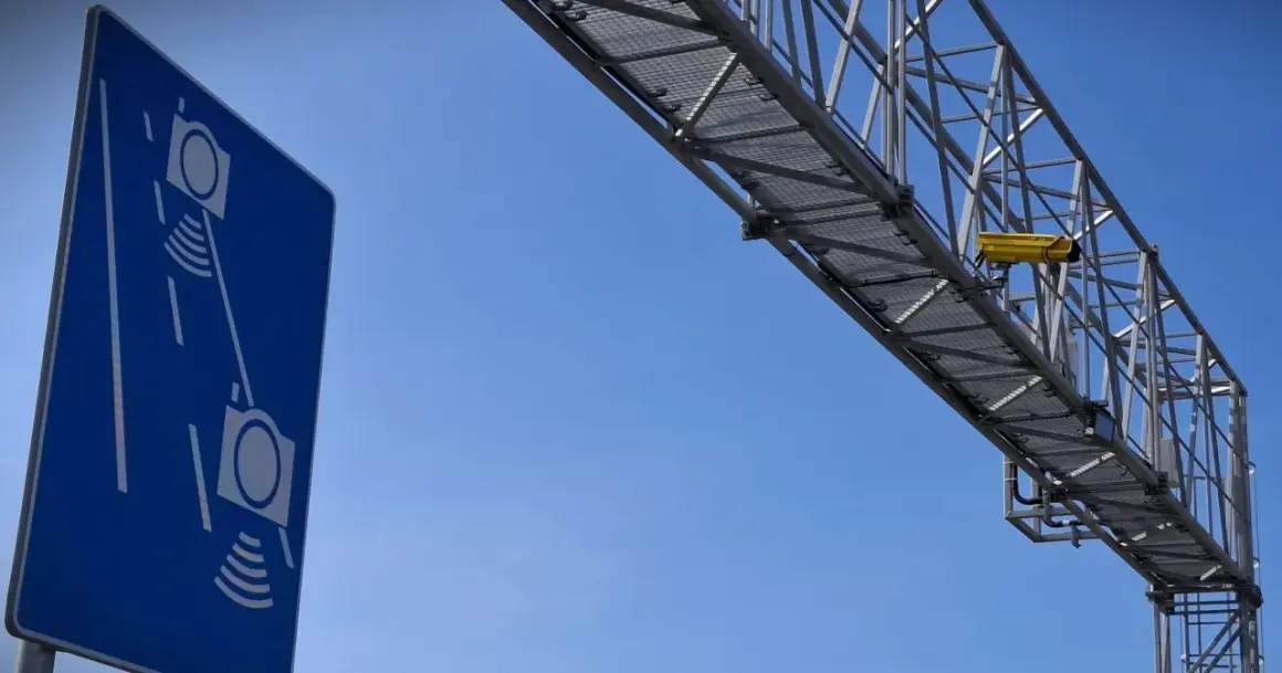 Niebieski znak informuje o odcinkowym pomiarze prędkości S7. Żółta kamera zamontowana na konstrukcji nad drogą rejestruje pojazdy.