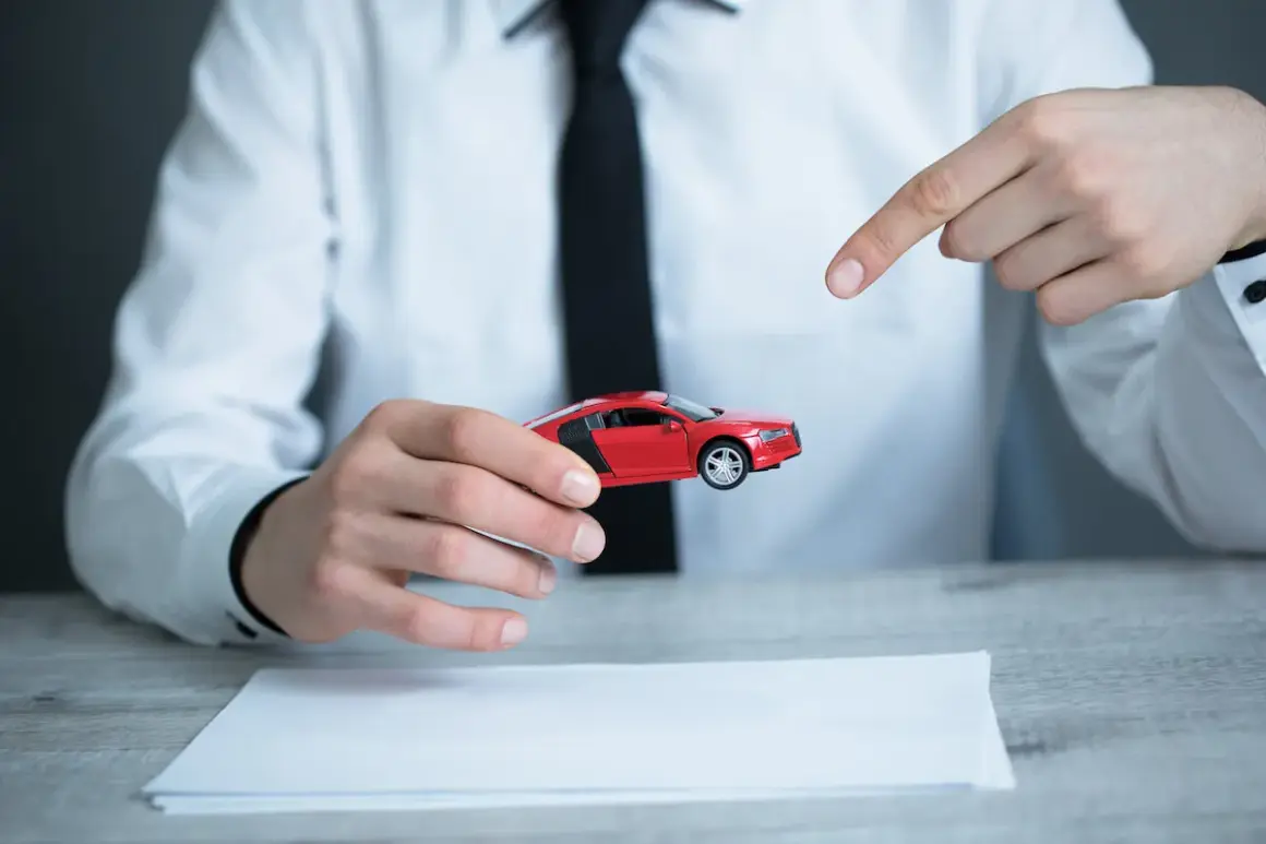 Mężczyzna w białej koszuli i czarnej krawacie prezentuje czerwony model samochodu, wskazując na niego palcem. Obok leży dokument, sugerujący umowę kupna sprzedaży samochodu z wyłączeniem rękojmi.